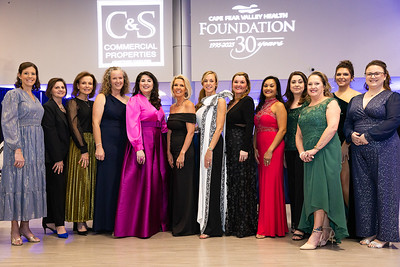 A group of women in elegant dresses poses together at an event, with a backdrop featuring a logo for a health foundation.